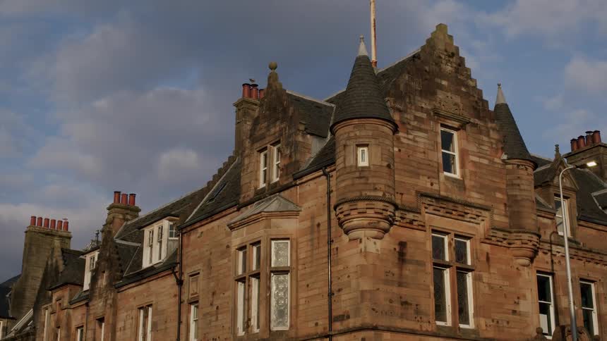 A classic stone building with intricate architecture and turrets is illuminated by the warm light of a setting sun under a partly cloudy sky