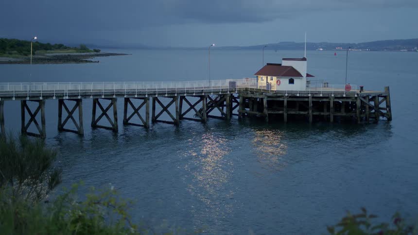 A quiet coastal pier with a lit building stands over calm water during a moody twilight evening