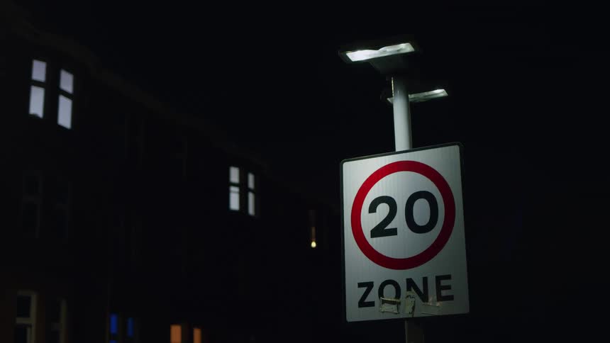 A 20 ZONE speed limit sign is illuminated by a streetlight at night in a quiet urban neighborhood