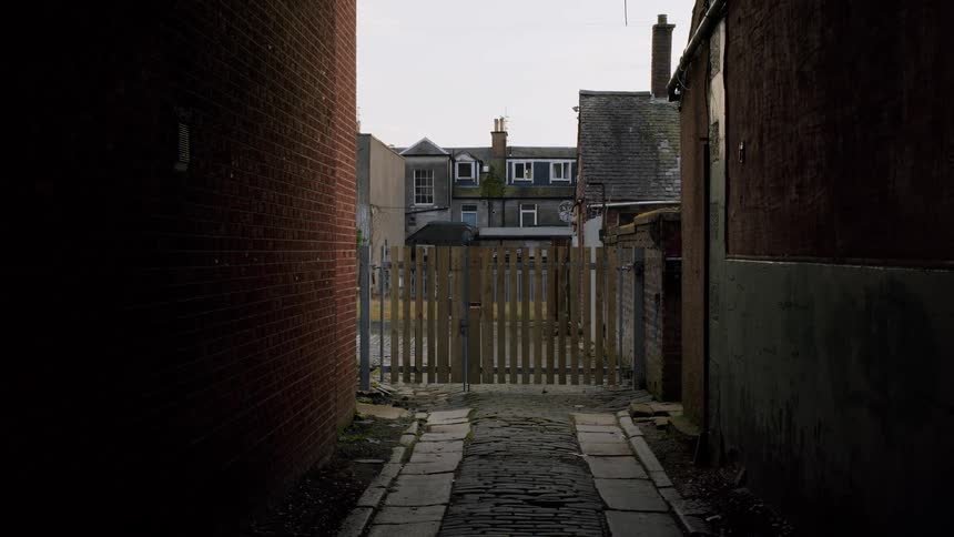 A dark and gritty urban cobblestone back alley leads to a wooden gate between two old brick buildings