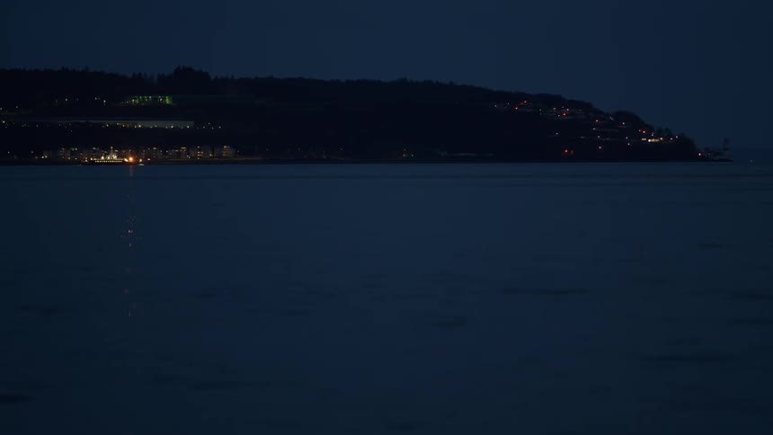 A serene view of calm ocean waters at night with the lights of a distant coastal town reflecting on the surface and a boat passing