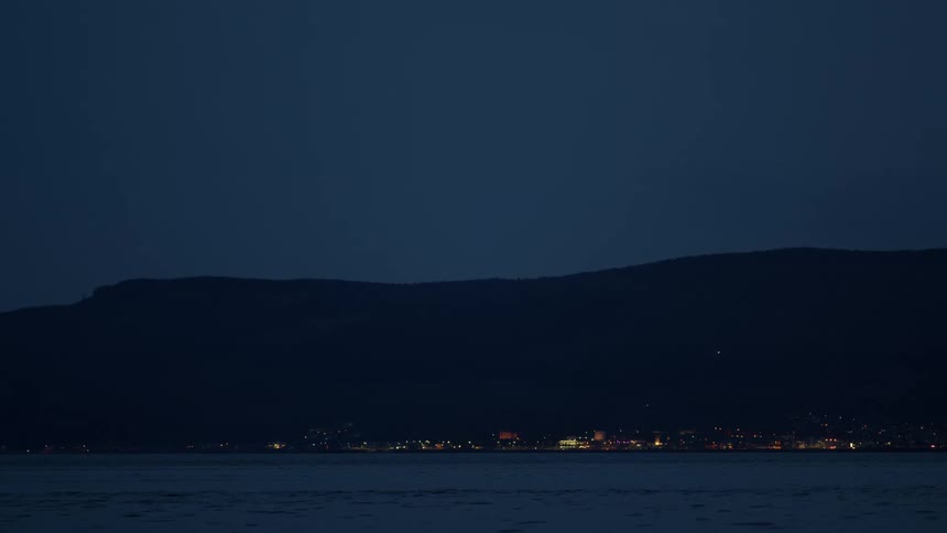 The lights of a small town glow across a calm lake at the base of a silhouetted mountain at night
