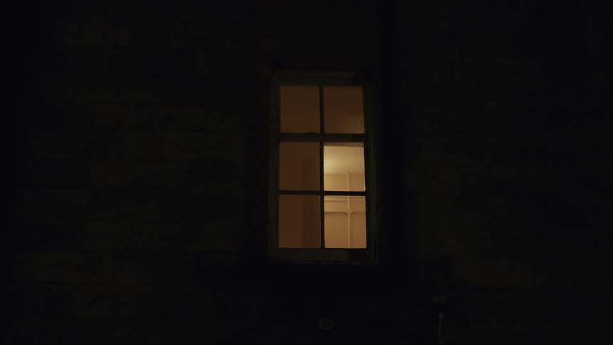 Handheld shot of a single warmly lit window seen from the exterior of a building at night