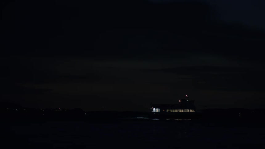 A boat with its lights on sails across the water at night, with a distant city skyline visible on the horizon
