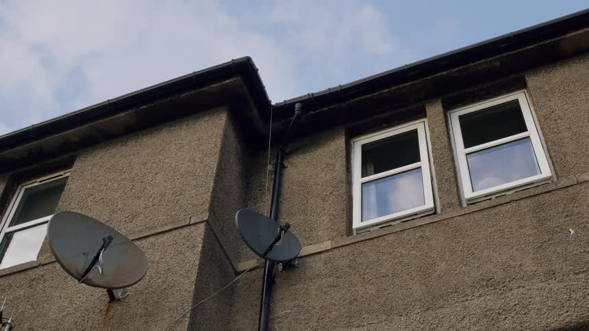 A low angle static shot of a gritty, pebble-dashed urban building with satellite dishes as clouds reflect in the windows