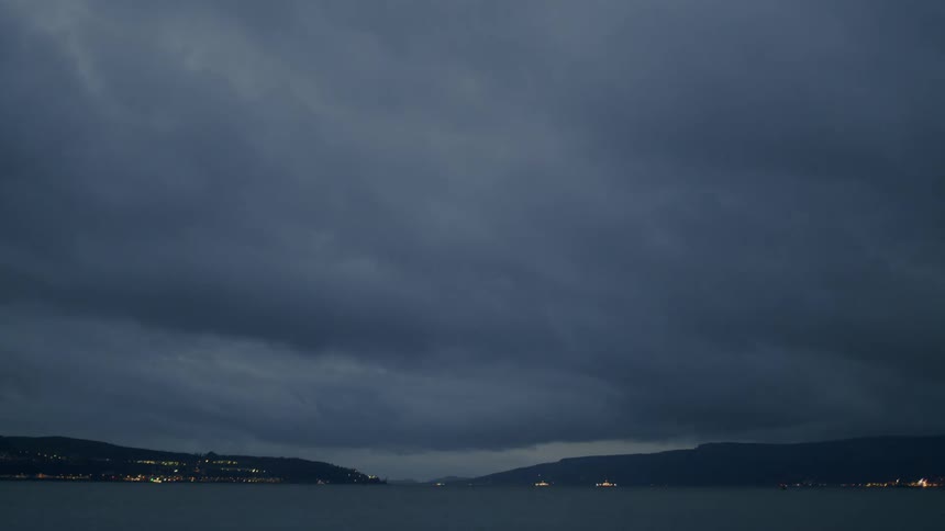 A dramatic time-lapse of heavy, dark storm clouds rolling over a body of water as day turns to night over the illuminated shores