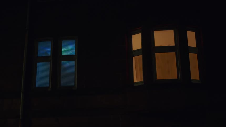 A house at night shows two contrasting illuminated windows, one with a steady warm glow and the other with shifting colored lights