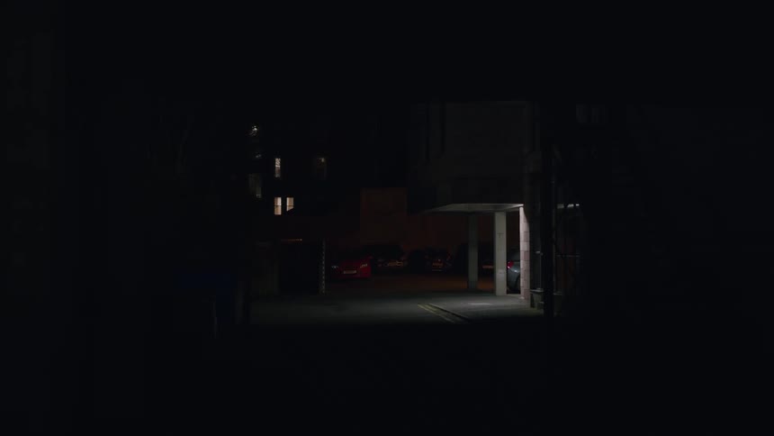 Static shot of a dark alleyway at night, revealing a single light illuminating a concrete pillar in a deserted parking lot