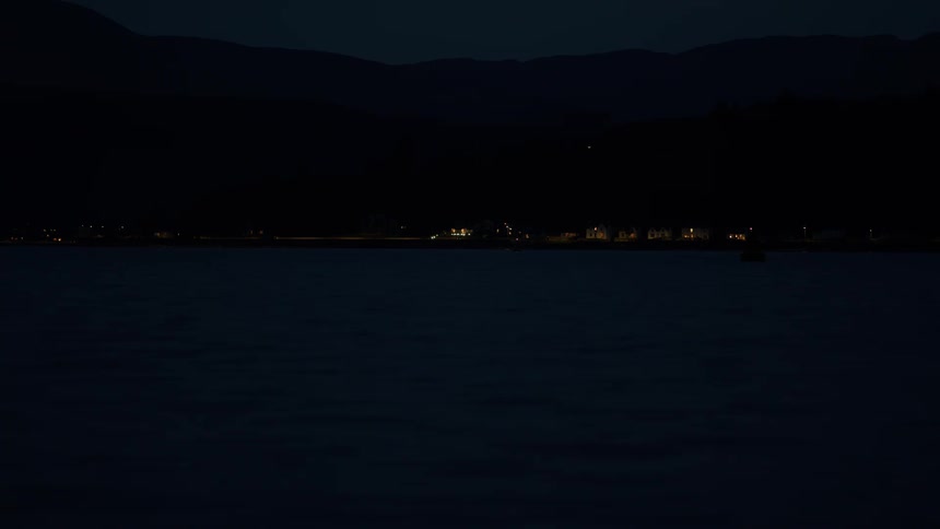 A tranquil night scene shows a calm sea with distant town lights on the shore and a flashing green navigational buoy