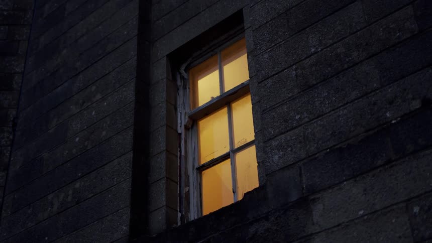 A warm light glows from a dilapidated window in a dark stone building at night