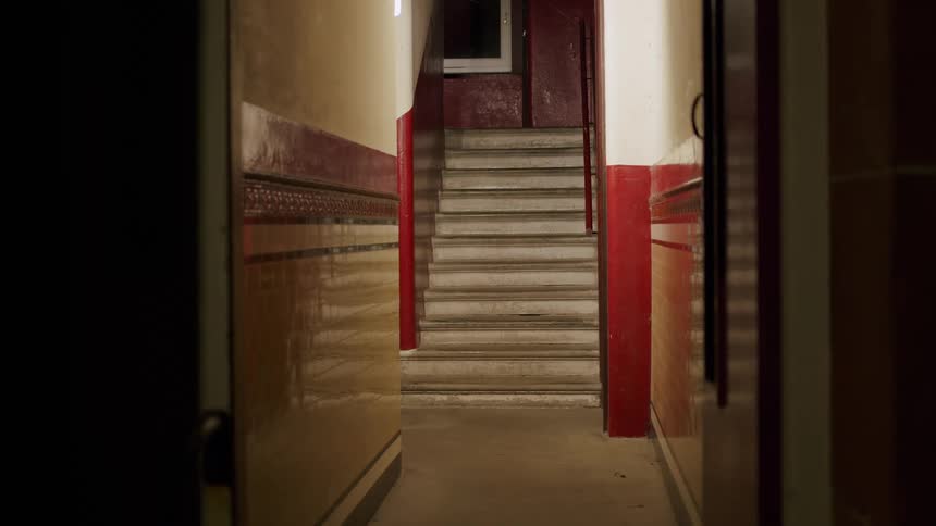 A static shot of a spooky and dimly lit hallway leading to a flight of stairs in an old building