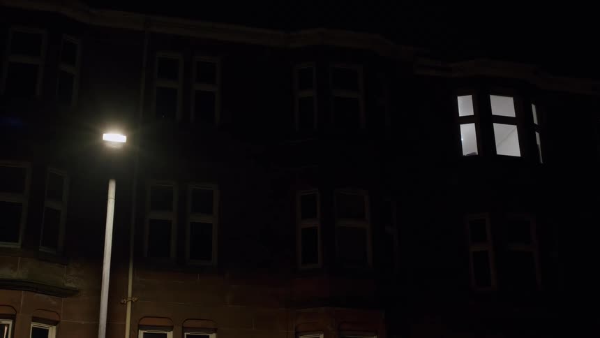 A single illuminated window on a dark apartment building at night with a street lamp shining nearby