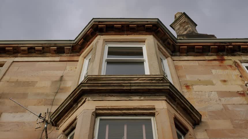 A low-angle, static shot shows the weathered facade of a traditional stone apartment building with a bay window under a grey, overcast sky