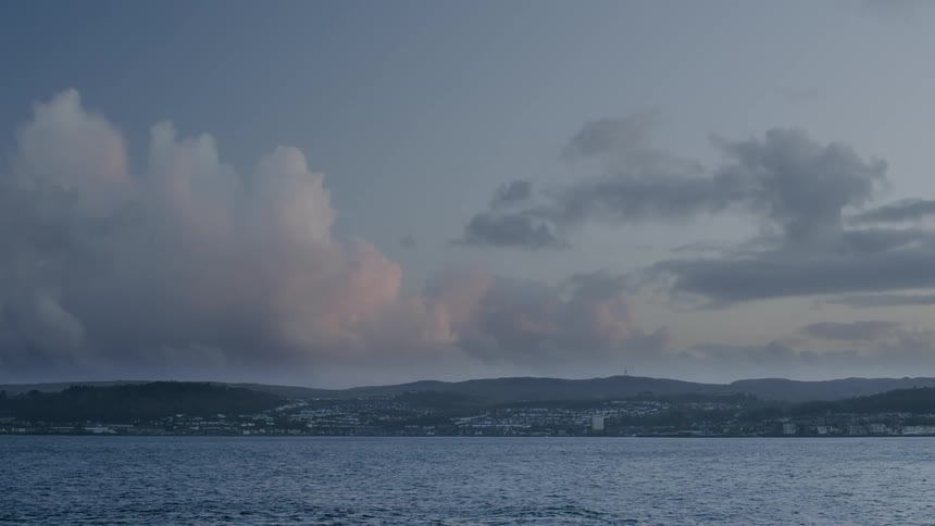 A serene wide shot of a coastal town on rolling hills across the water under a dramatic cloudy sky at dusk