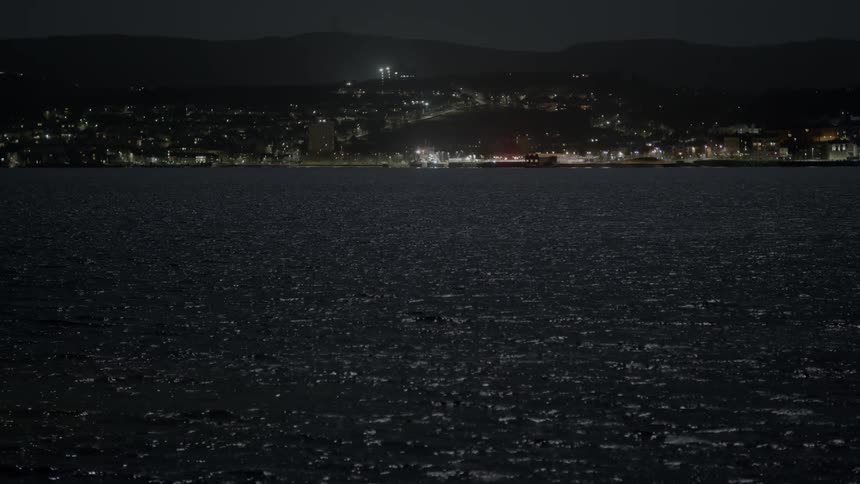 The dark waters of the sea gently ripple at night, with a distant coastal city illuminated on the horizon