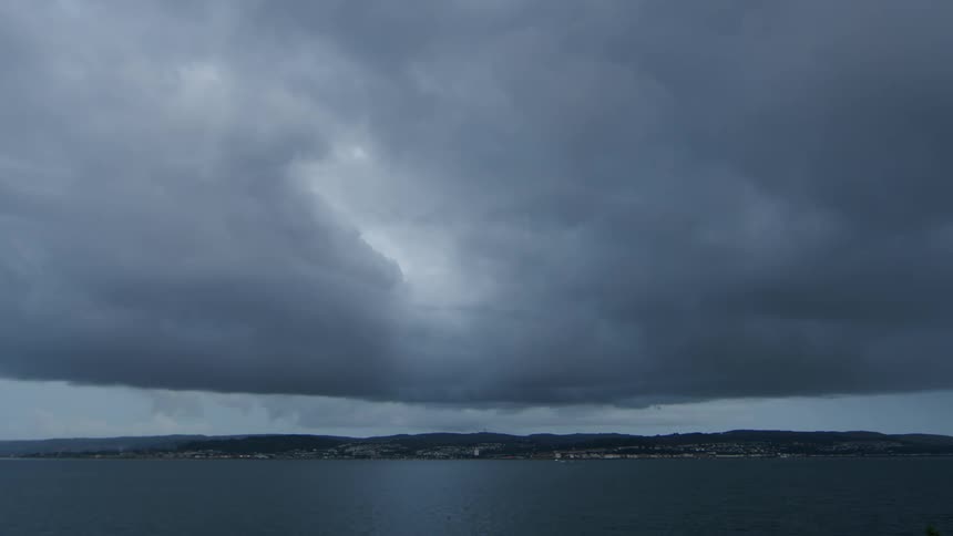 Ominous gray storm clouds hang low over a calm ocean, with a distant coastal town on the horizon