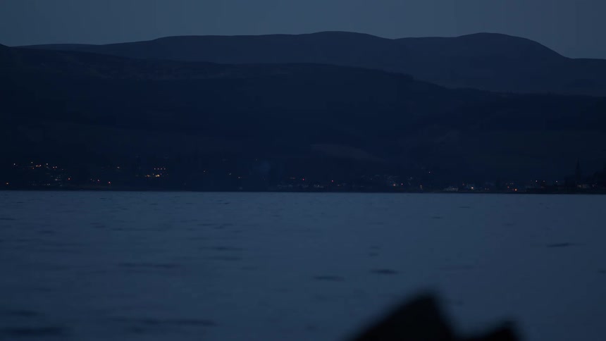 A serene view across calm water towards the twinkling lights of a distant town nestled at the base of mountains at dusk