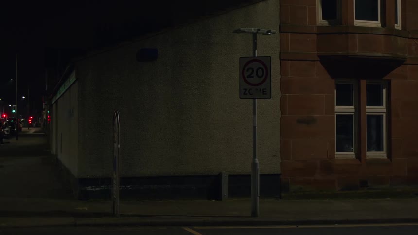 A faulty flashing 20 mph zone street sign stands on a dark, empty city corner at night