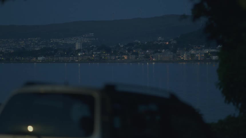 A distant coastal city at night, viewed from across the water with a car in the foreground