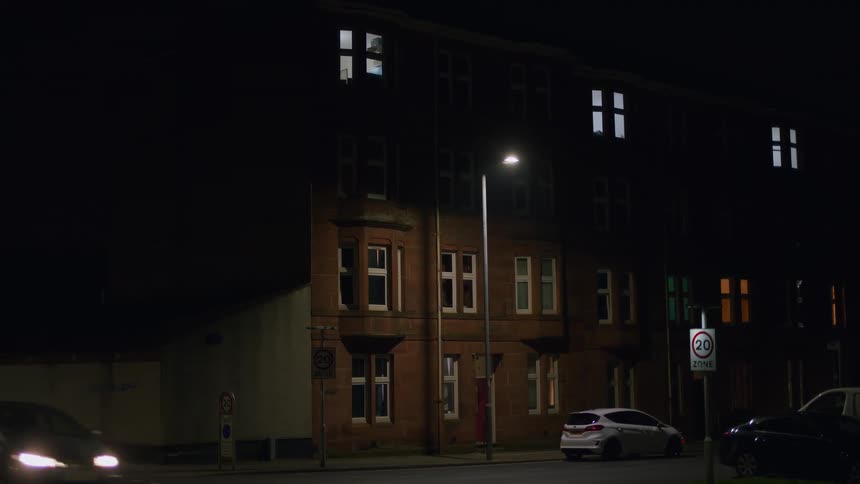 A static shot of a residential city street at night, with a single streetlamp illuminating a brick apartment building as a car drives by