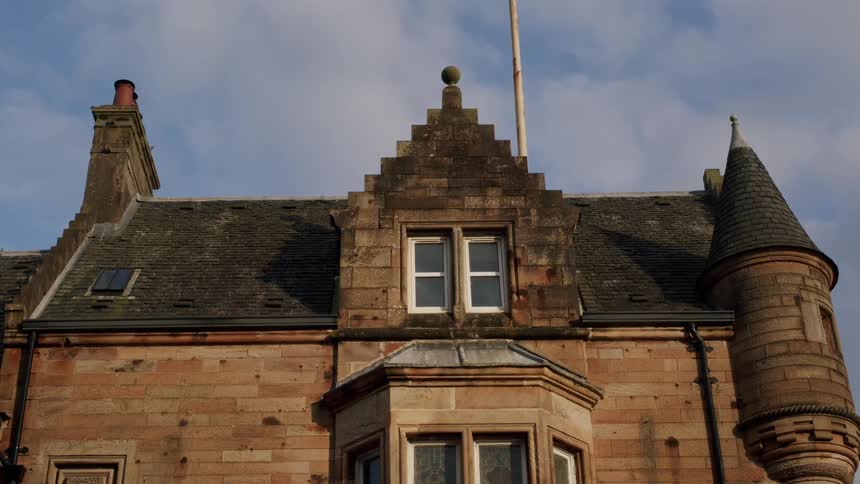 A low-angle static shot shows the roofline of a traditional stone building with a turret and chimney