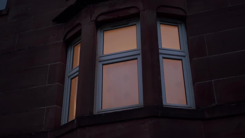 A warm orange light glows from the window of a dark stone building at night, creating a cozy and inviting scene