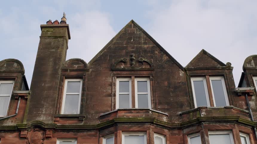 A static low-angle shot of the top floor of a weathered, old red sandstone building against an overcast sky