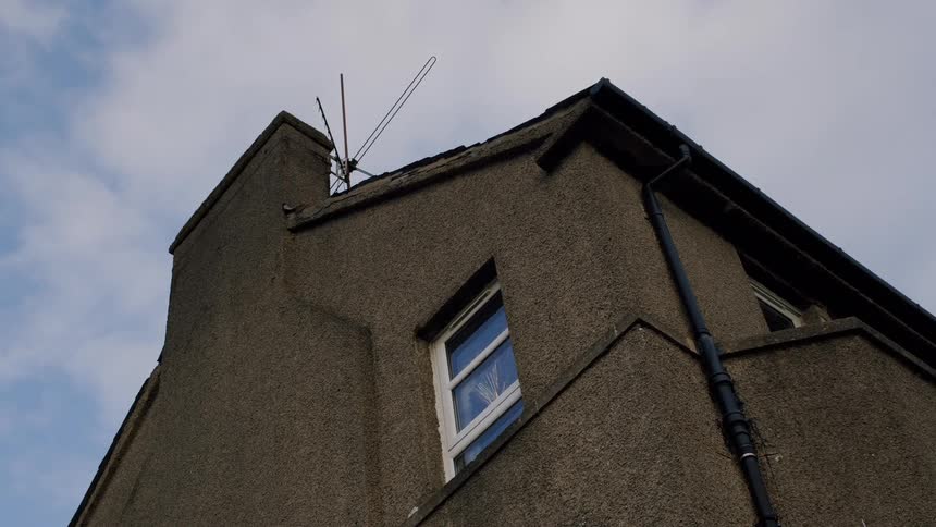 A static low-angle shot of a pebbledash house with an old-fashioned TV aerial on the roof against a cloudy blue sky