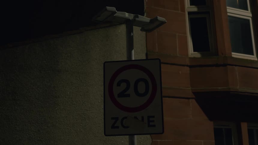 A malfunctioning street light flickers on and off, illuminating a 20 mile-per-hour zone sign on a dark city street at night