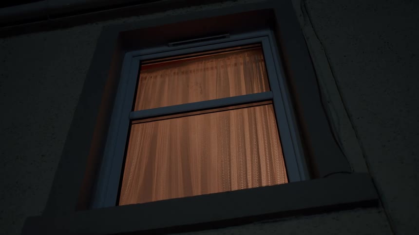 Warm light glows from behind the curtains of a house window on a dark and quiet night