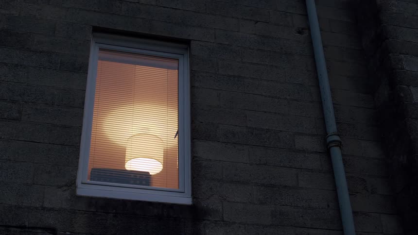 A warm lamp glows through a window with blinds on a dark stone building at night