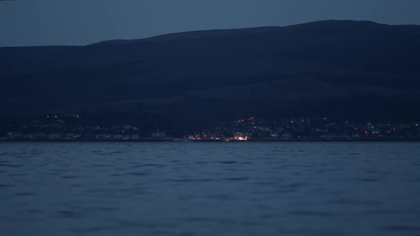 A quiet coastal village is illuminated by streetlights and homes at night, viewed from across the calm water