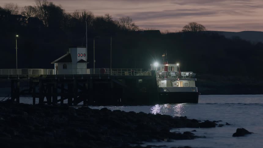 A passenger ferry sits docked at a quiet rural pier at dusk, its lights reflecting on the still water