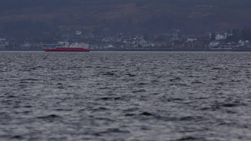 A bright red ferry ship sails on a gray sea with a coastal town and rolling hills in the background on an overcast day