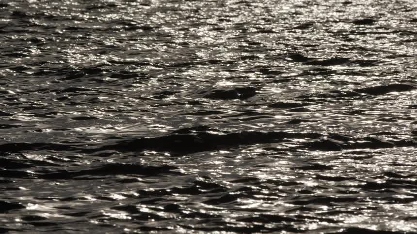 Sunlight sparkles and reflects off the dark surface of gently rippling water in slow motion