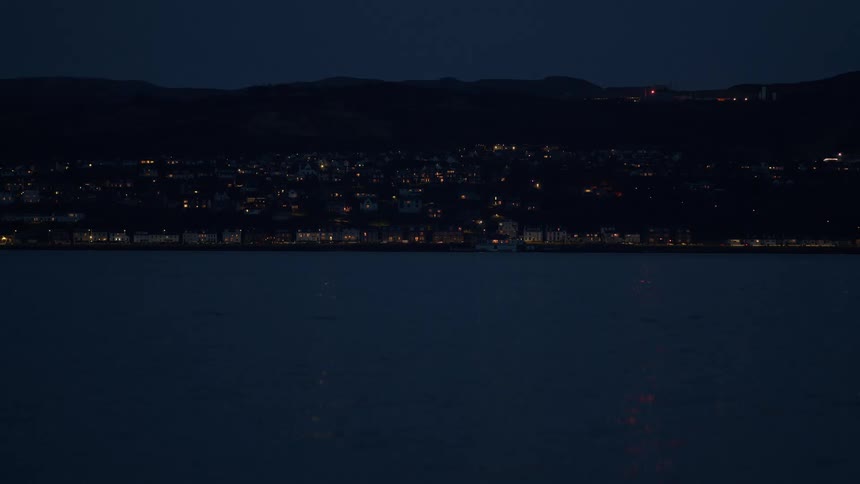 A wide shot of a small coastal town illuminated at night, viewed from across the dark, serene water