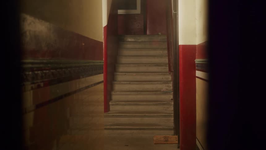 An eerie dolly out shot reveals a dark and dirty hallway in an abandoned building with a staircase in the distance