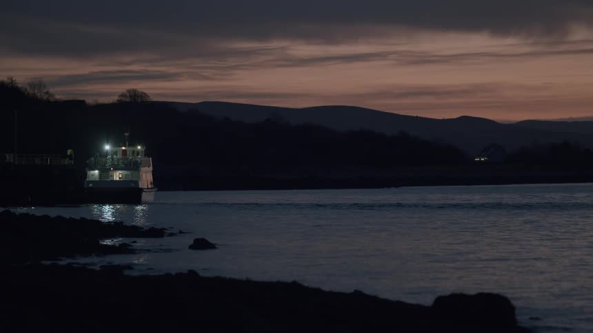 A lit ferry boat sits docked at a pier on calm water against a silhouetted landscape at twilight