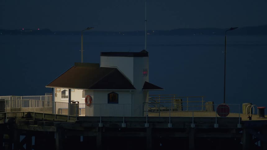 A quiet, lonely white building sits on a wooden pier over the dark water at dusk