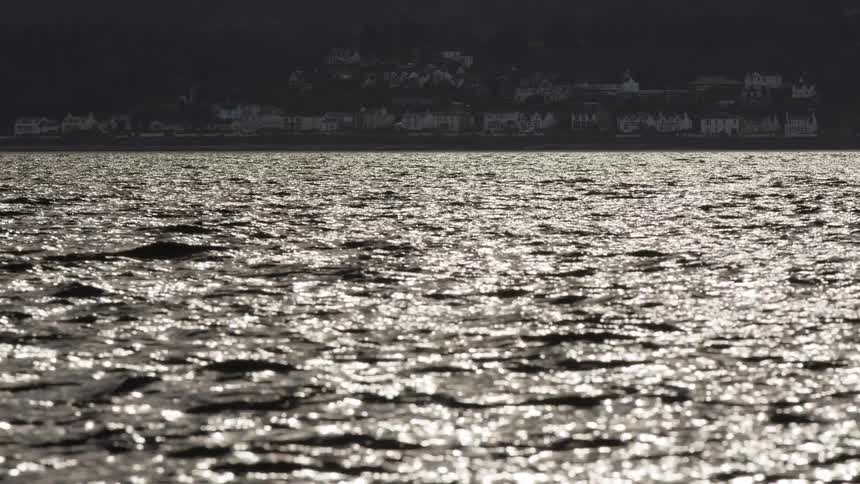 Sunlight glitters on the dark, rippling water surface with a silhouetted coastal town in the distance