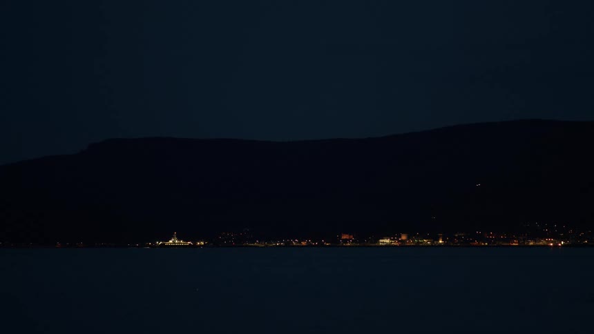 Distant city and ship lights glow across the water against a dark mountain silhouette at night