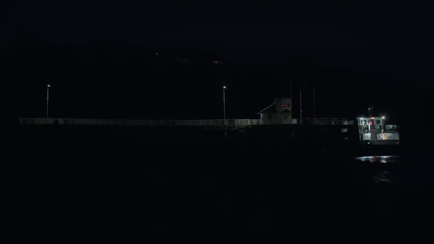 A lone ship is docked at an illuminated pier in the middle of a very dark night