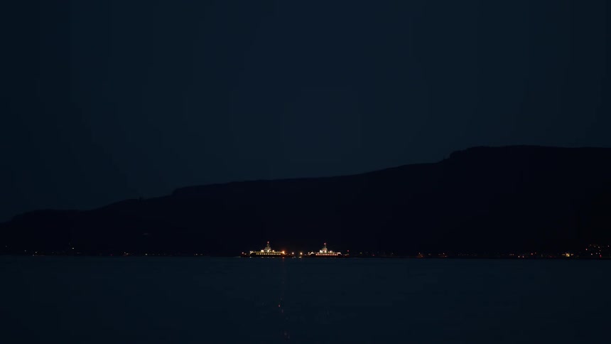 A tranquil nighttime shot captures the glowing lights of two ferry boats passing in front of a mountainous landscape