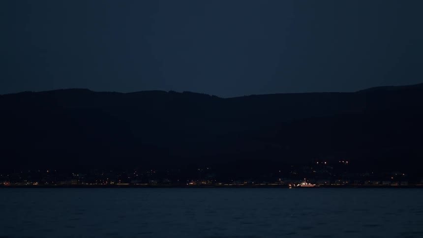 A boat sails across the bay at night, with the lights of a distant coastal town twinkling on the horizon