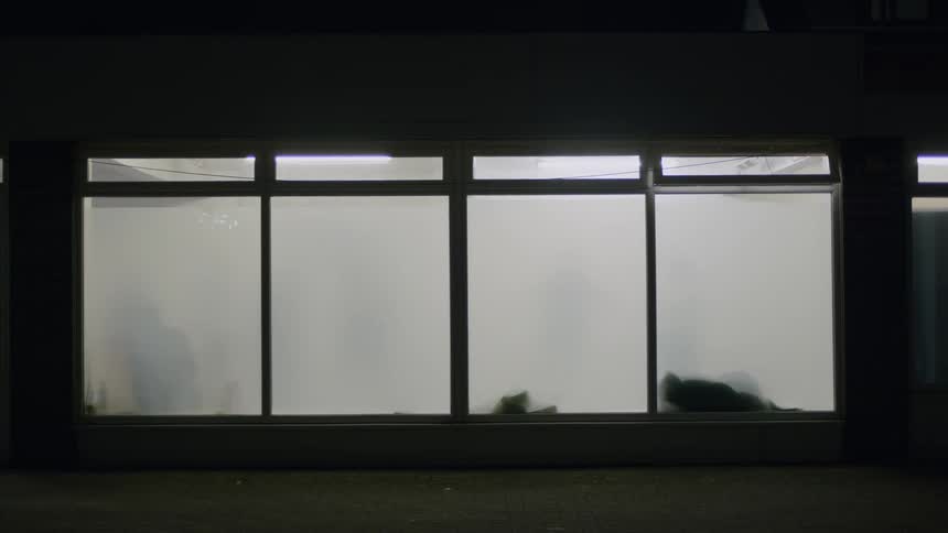 Mysterious human silhouettes are seen through a large, brightly lit, frosted window at night