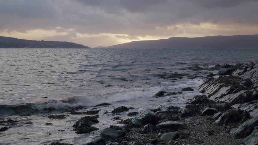 Slow-motion waves gently wash over a rocky, seaweed-strewn shore under a dramatic, cloudy sky at dusk