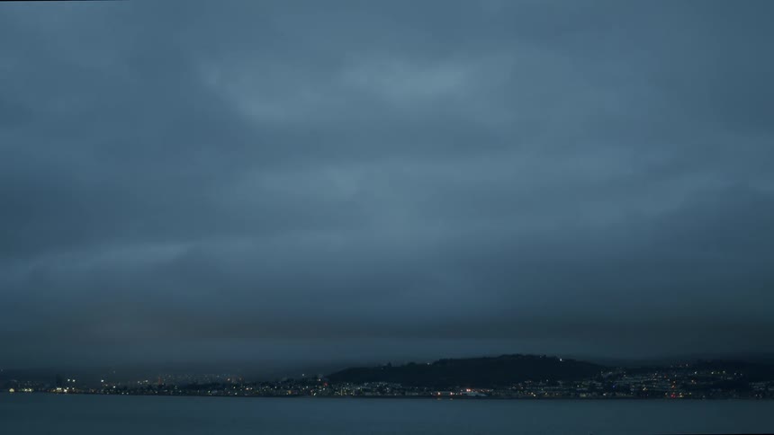 A coastal city is shrouded in dark, moody clouds and fog at twilight, with city lights twinkling in the distance