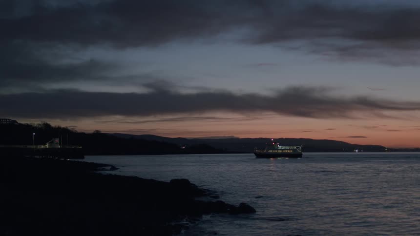 A ferry boat sails across the water near a rocky coastline as the last light of sunset fades into dusk