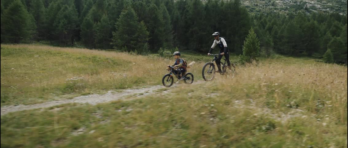 A father and son mountain bike with their dog running alongside them on a scenic alpine trail in slow motion