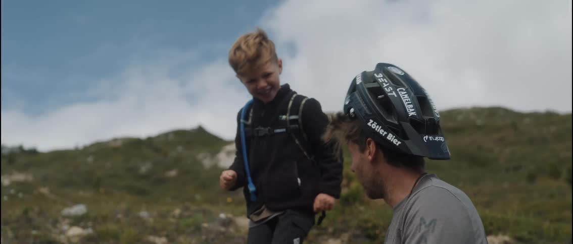 A happy young boy playfully puts a bicycle helmet on his smiling father's head as they take a break in the mountains